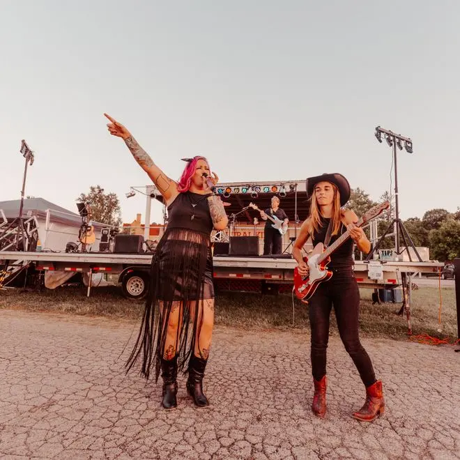 Indy Annies performing outdoors, with vocalist Breezey Fox and guitarist Krista Hoose in front of the stage.
