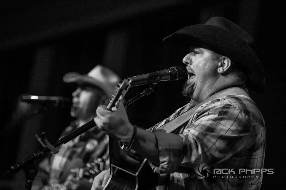 Joe Hess singing and playing acoustic guitar, with another musician behind him; original Rick Phipps Photography credit remains visible.